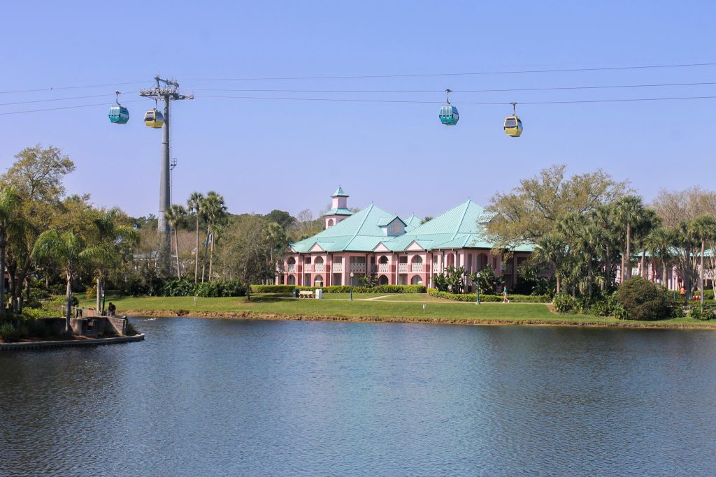 Disney's Caribbean Beach Resort - Disney Moderate Resort. Pastel pink resort buildings with tea roofs sit along a waterfront at Disney's Caribbean Beach Resort while Skyliner gondolas glide overhead in a clear blue sky.