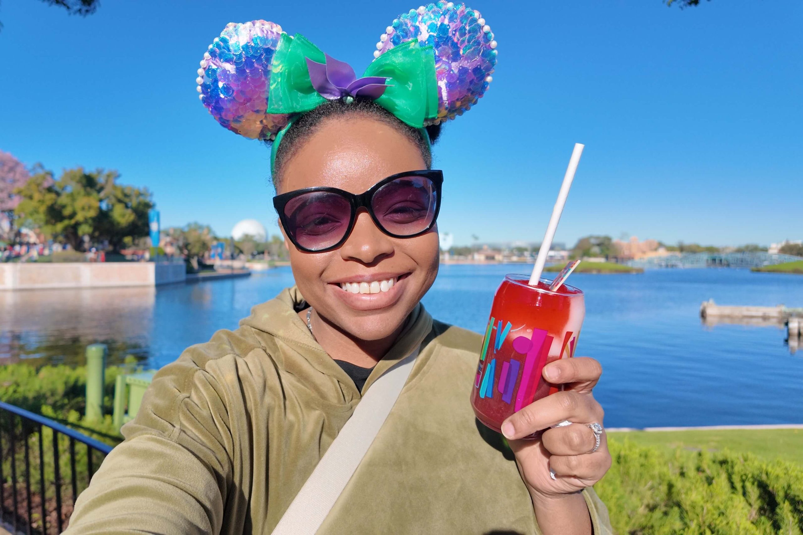 Woman wearing sparkling purple and teal Minnie ears and oversized sunglasses smiles while holding a frozen red drink beside the World Showcase Lagoon at EPCOT Disney World. Sipping cold drinks and taking relaxing breaks by the water are simple ways to stay cool at Disney World on a sunny park day.