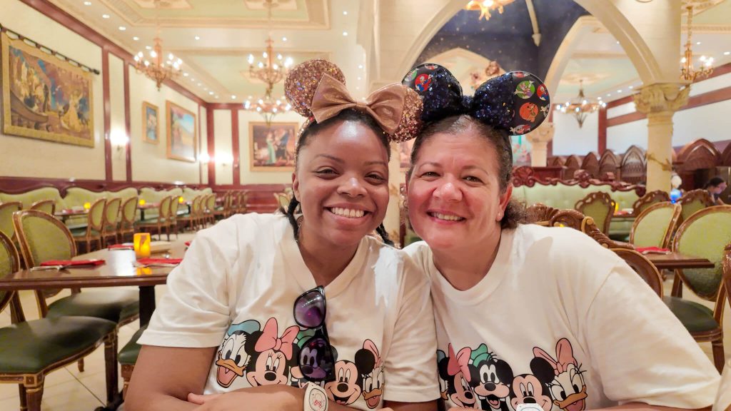 Two Disney moms smiling wearing matching Disney character T shirts and sparkly Minnie ears sit together inside Be Our Guest, a castle themed restaurant at Walt Disney World. The warm elegant dining room creates a celebratory atmosphere that fits a girls trip and offers inspiration alongside mom tips for disney world.