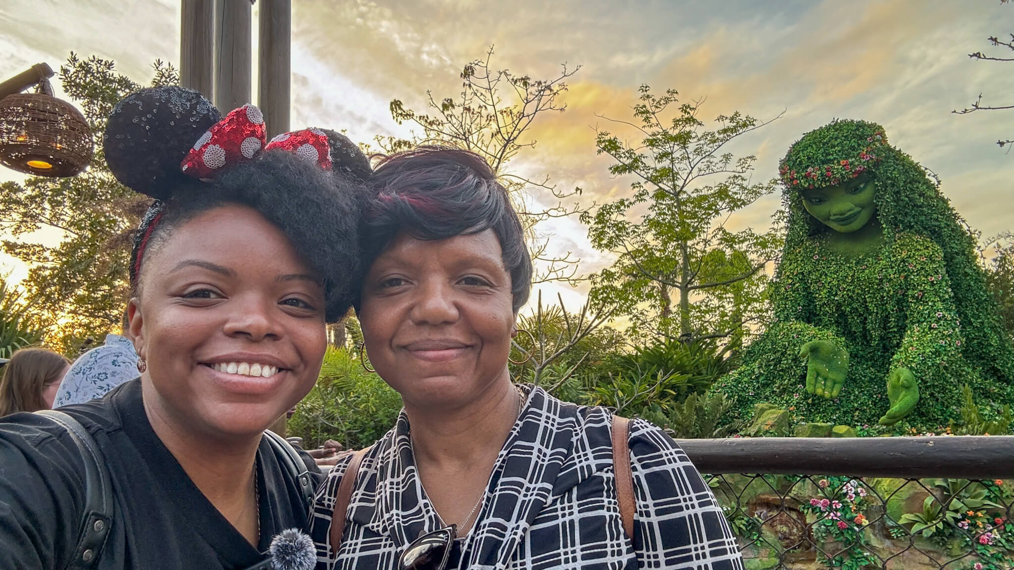 Things to do before a Disney World trip - 30 days to go countdown. Two women (mother and daughter) smile for a selfie at Walt Disney World with a large green floral character behind them and lush tropical landscaping surrounding the area.